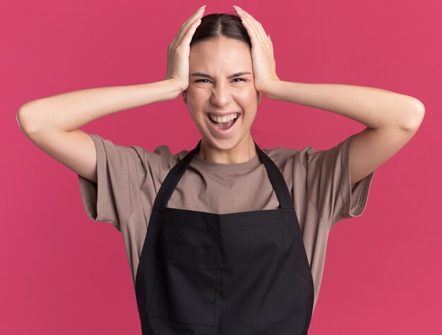 Joyeuse jeune fille de coiffeur brune en uniforme met les mains sur la tête isolée sur un mur rose avec espace copie