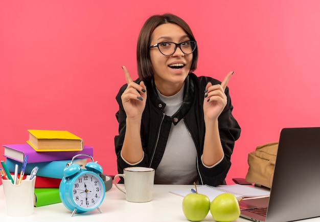 Joyeuse jeune fille étudiante portant des lunettes assis au bureau à faire ses devoirs en levant les doigts isolés sur rose