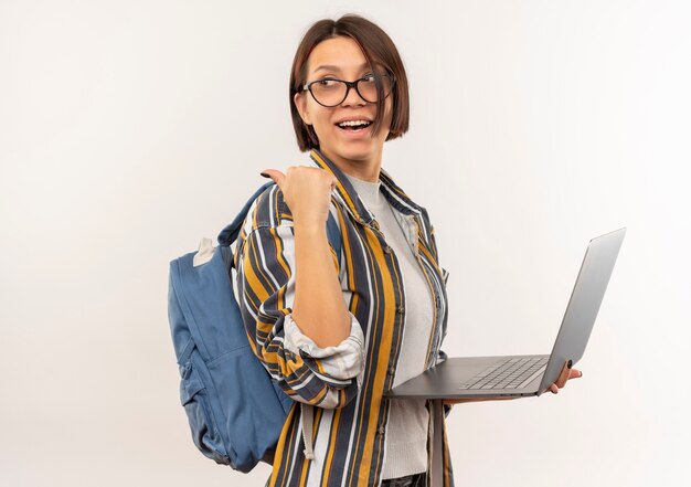 Joyeuse jeune fille étudiante portant des lunettes et sac à dos debout en vue de profil tenant un ordinateur portable pointant derrière isolé sur blanc