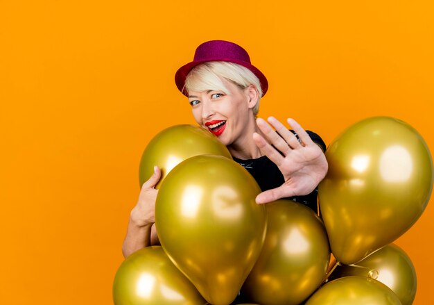 Joyeuse jeune fille de fête blonde portant un chapeau de fête debout derrière des ballons saisissant l'un d'entre eux faisant le geste d'arrêt à la caméra en regardant la caméra isolée sur fond orange