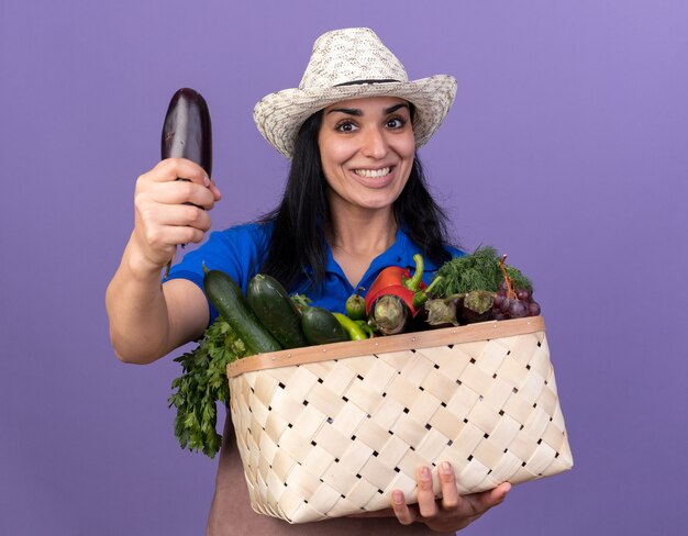 Joyeuse jeune fille de jardinier caucasien en uniforme et chapeau tenant un panier de légumes étirant l'aubergine vers la caméra isolée sur un mur violet