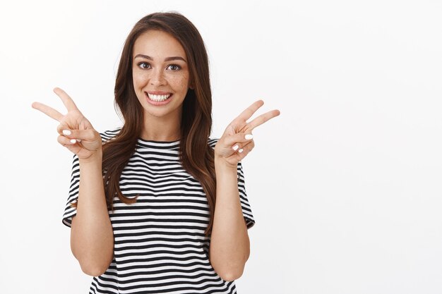 Joyeuse jeune fille optimiste, mignonne et drôle avec des taches de rousseur en t-shirt rayé partageant la positivité, souriant amicalement et montrant un signe de paix ou de victoire de bonne volonté, debout sur un mur blanc sans soucis