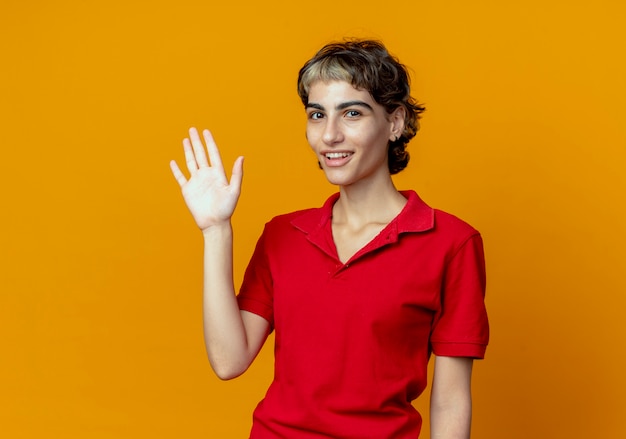 Joyeuse jeune fille de race blanche avec coupe de cheveux de lutin faisant le geste salut à la caméra isolée sur fond orange avec espace de copie