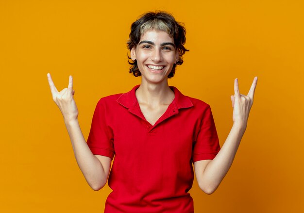 Joyeuse jeune fille de race blanche avec coupe de cheveux de lutin faisant des signes de roche isolés sur fond orange