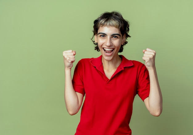 Photo gratuite joyeuse jeune fille de race blanche avec coupe de cheveux de lutin serrant les poings isolés sur fond vert olive avec espace de copie