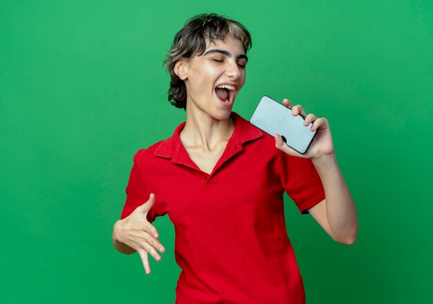 Joyeuse jeune fille de race blanche avec coupe de cheveux de lutin tenant un téléphone mobile faisant semblant de chanter à l'aide de téléphone comme microphone en gardant la main dans l'air avec les yeux fermés isolé sur fond vert avec espace copie