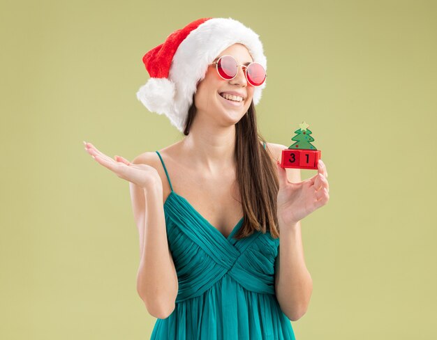 Joyeuse jeune fille de race blanche à lunettes de soleil avec bonnet de noel tenant ornement d'arbre de Noël et regardant à côté