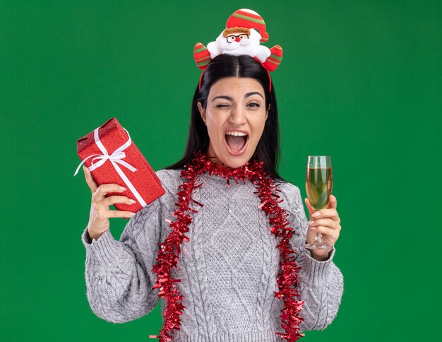 Joyeuse jeune fille de race blanche portant bandeau de père Noël et guirlande de guirlandes autour du cou tenant le paquet cadeau et verre de champagne un clin d'oeil isolé sur le mur vert