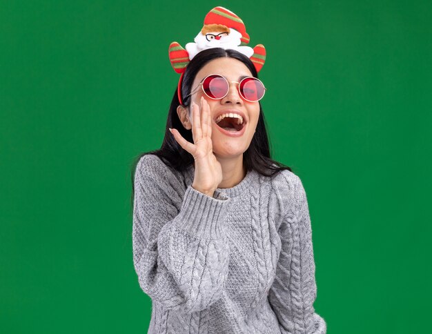 Joyeuse jeune fille de race blanche portant bandeau de père Noël avec des lunettes chuchotant isolé sur mur vert avec espace copie