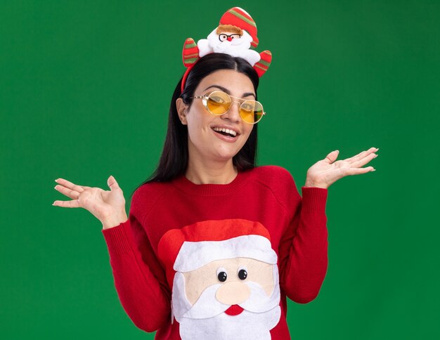 Joyeuse jeune fille de race blanche portant bandeau de père Noël et pull avec des lunettes regardant la caméra montrant les mains vides isolés sur fond vert