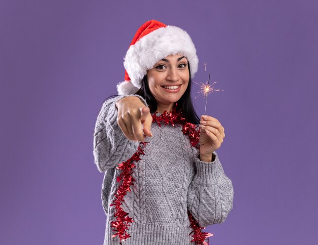 Joyeuse jeune fille de race blanche portant chapeau de Noël et guirlande de guirlandes autour du cou tenant un cierge magique isolé sur mur violet avec espace de copie