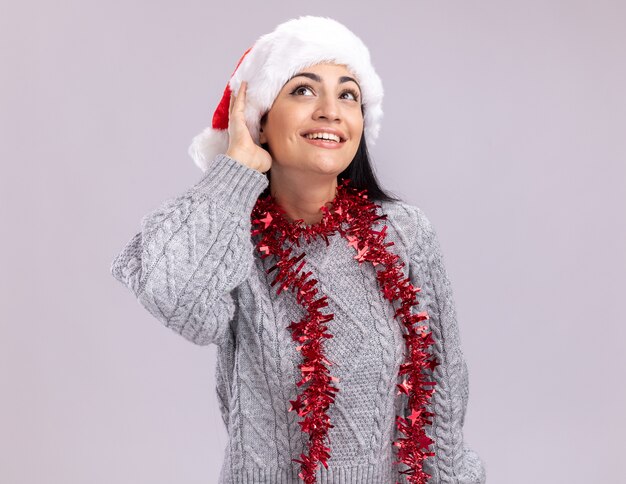 Joyeuse jeune fille de race blanche portant chapeau de Noël et guirlande de guirlandes autour du cou toucher hat en levant isolé sur fond blanc