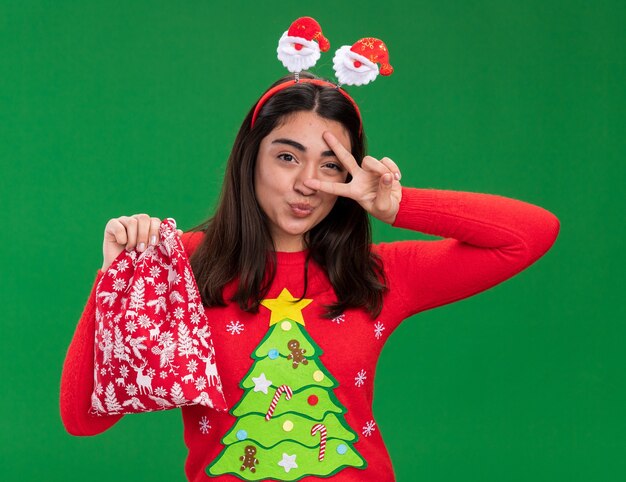 Joyeuse jeune fille de race blanche avec le signe de la victoire des gestes du bandeau de Santa et tient le sac de cadeau de Noël isolé sur fond vert avec espace de copie