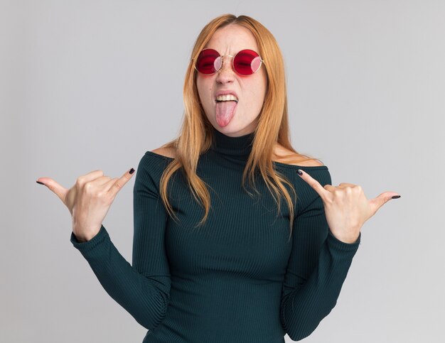 Joyeuse jeune fille rousse au gingembre avec des taches de rousseur dans des lunettes de soleil sort la langue faisant accrocher le geste lâche sur blanc