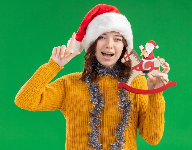 Joyeuse jeune fille slave avec bonnet de Noel et avec guirlande autour du cou tenant le père Noël sur la décoration de cheval à bascule et pointant vers le haut isolé sur fond vert avec espace copie