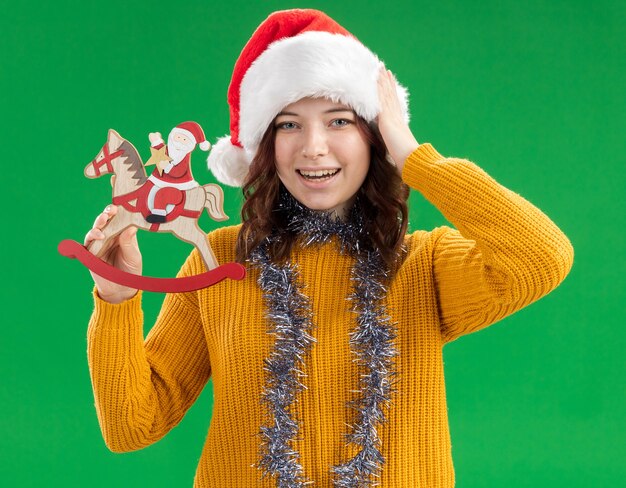 Joyeuse jeune fille slave avec bonnet de noel et guirlande autour du cou tient le père noël sur une décoration de cheval à bascule et met la main sur la tête isolée sur un mur vert avec espace de copie