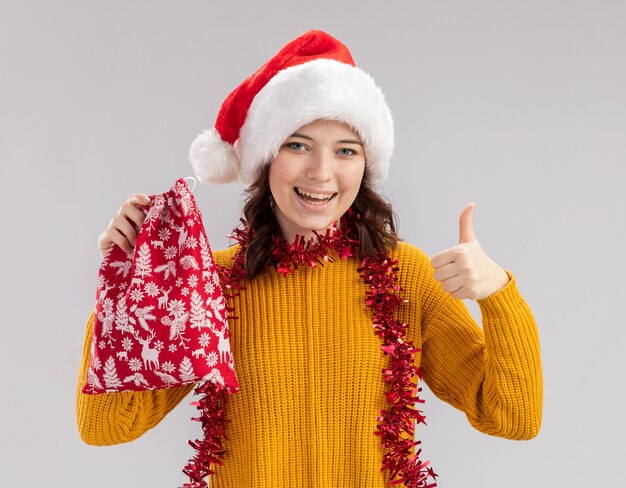 Joyeuse jeune fille slave avec bonnet de Noel et avec guirlande autour du cou tient le sac-cadeau de Noël et les pouces vers le haut isolé sur fond blanc avec espace copie