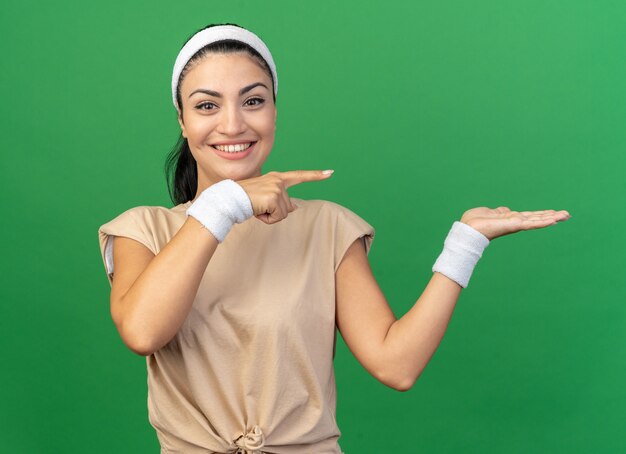 Joyeuse jeune fille sportive caucasienne portant un bandeau et des bracelets montrant une main vide pointant sur le côté isolé sur un mur vert