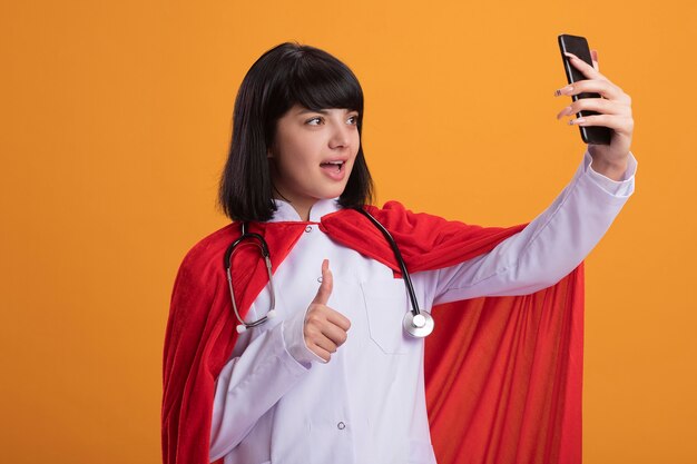 Joyeuse jeune fille de super-héros portant un stéthoscope avec une robe médicale et une cape prendre un pouce selfie isolé sur orange