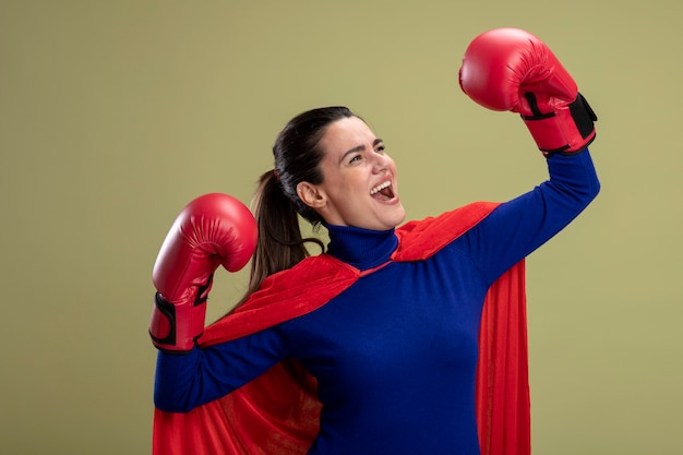 Joyeuse jeune fille de super-héros regardant côté portant des gants de boxe montrant un geste fort isolé sur vert olive