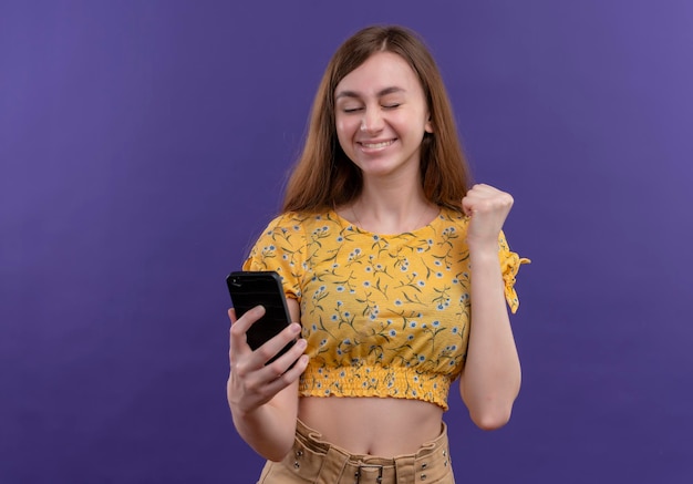 Joyeuse jeune fille tenant un téléphone mobile et levant le poing sur un espace violet isolé avec copie espace