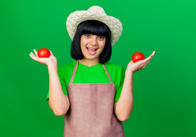 Joyeuse jeune jardinière en uniforme portant un chapeau de jardinage détient des tomates isolées