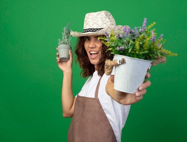 Photo gratuite joyeuse jeune jardinière en uniforme portant chapeau de jardinage tenant des fleurs dans des pots de fleurs