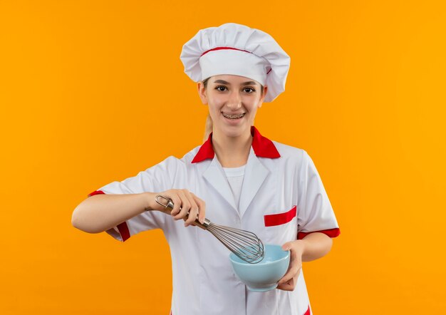 Joyeuse jeune jolie cuisinière en uniforme de chef avec appareil dentaire tenant un fouet et un bol isolé sur l'espace orange