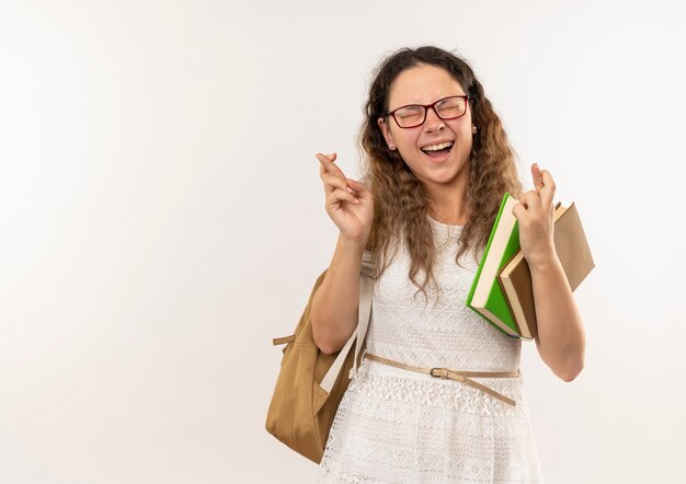 Joyeuse jeune jolie écolière portant des lunettes et sac à dos tenant des livres et croisant les doigts avec les yeux fermés isolés sur blanc