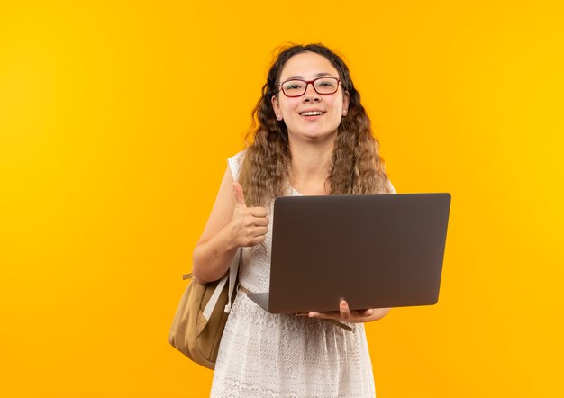Joyeuse jeune jolie écolière portant des lunettes et sac à dos tenant un ordinateur portable montrant le pouce vers le haut isolé sur jaune