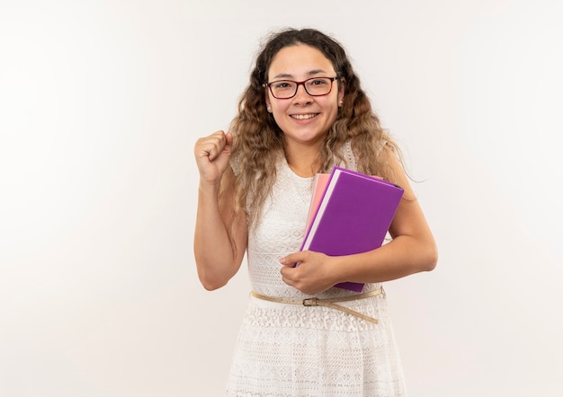 Joyeuse jeune jolie écolière portant des lunettes tenant des livres levant le poing isolé sur blanc