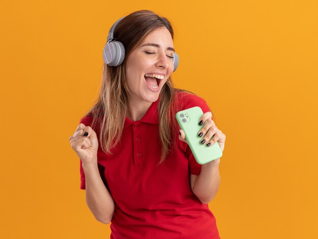 Joyeuse jeune jolie femme sur les écouteurs garde le poing et tient le téléphone isolé sur un mur orange