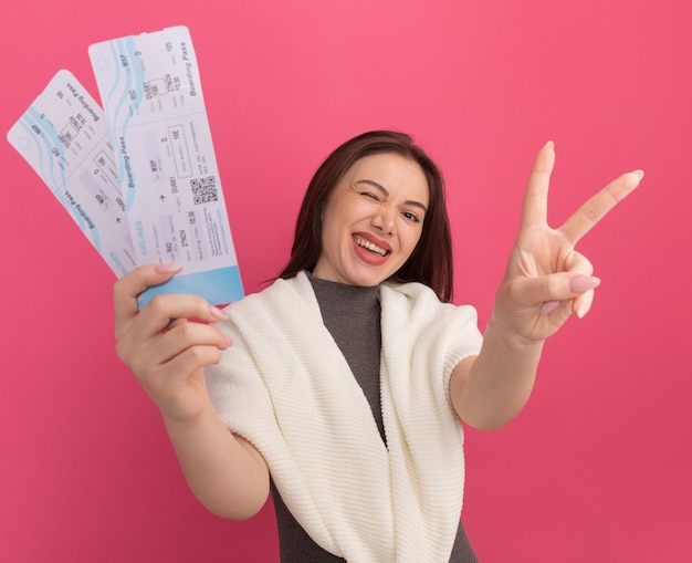 Joyeuse jeune jolie femme étirant des billets d'avion faisant un signe de paix isolé sur un mur rose