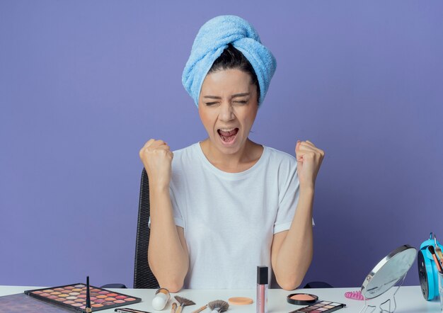Joyeuse jeune jolie fille assise à la table de maquillage avec des outils de maquillage et avec une serviette de bain sur la tête en serrant les poings avec les yeux fermés isolé sur fond violet