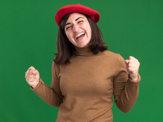 Joyeuse jeune jolie fille caucasienne avec chapeau de béret garde les poings isolés sur un mur vert avec espace de copie