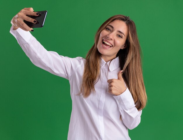 Joyeuse jeune jolie fille caucasienne pouces vers le haut et tient le téléphone isolé sur un mur vert avec espace de copie