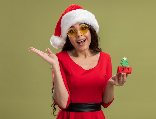 Joyeuse jeune jolie fille portant bonnet de Noel et lunettes tenant jouet arbre de Noël avec date à la recherche montrant la main vide
