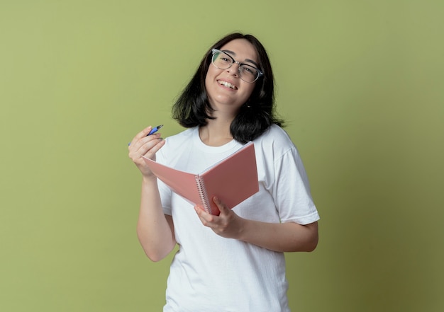 Joyeuse jeune jolie fille portant des lunettes tenant un stylo et un bloc-notes à tout droit