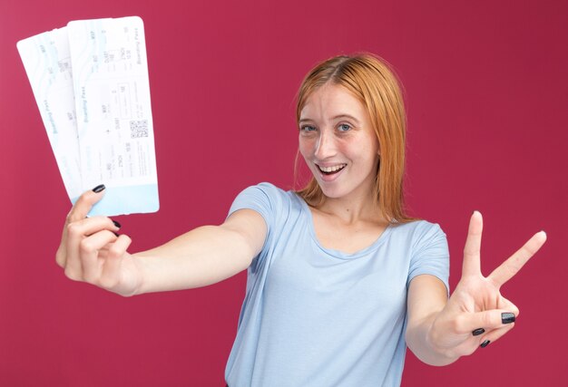 Joyeuse jeune rousse au gingembre avec des taches de rousseur gesticulant le signe de la victoire et tenant des billets d'avion isolés sur un mur rose avec espace pour copie