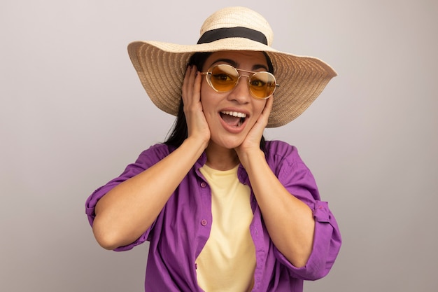 Joyeuse jolie femme brune à lunettes de soleil avec chapeau de plage met les mains sur le visage isolé sur mur blanc