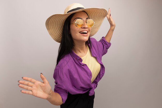 Joyeuse jolie femme brune à lunettes de soleil avec chapeau de plage se dresse avec les mains levées à l'avant isolé sur mur blanc