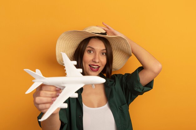 Joyeuse jolie femme caucasienne avec chapeau de plage détient avion modèle sur orange