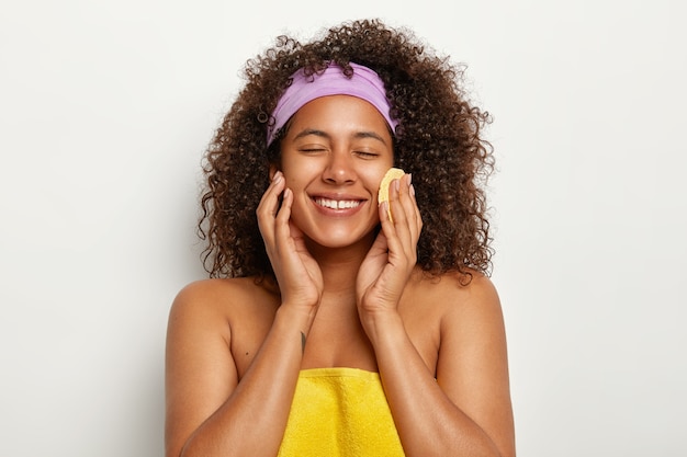 joyeuse jolie femme avec coupe de cheveux afro, essuie le visage avec une éponge cosmétique ronde, enlève le maquillage, a une beauté naturelle, un bandeau weras