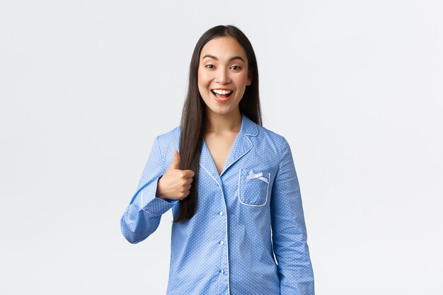 Joyeuse jolie fille asiatique en pyjama bleu souriante heureuse et montrant le pouce levé en signe d'approbation ou similaire, recommande un produit, de grande qualité, montrant du bon travail ou du bon travail, fond blanc.