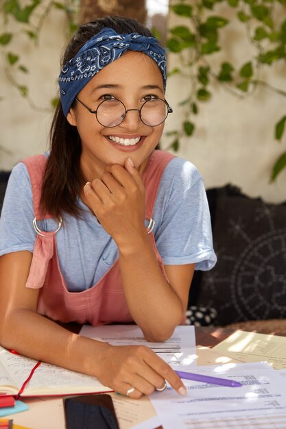 Joyeuse jolie fille métisse a un sourire charmant parfait, montre des dents blanches, porte un bandeau bleu sur la tête, occupée à écrire les notes nécessaires