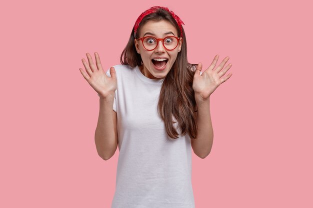 Joyeuse jolie fille s'amuse, fait des gestes actifs, rit de quelque chose de drôle, porte un bandeau rouge et un t-shirt décontracté, montre des paumes à la caméra