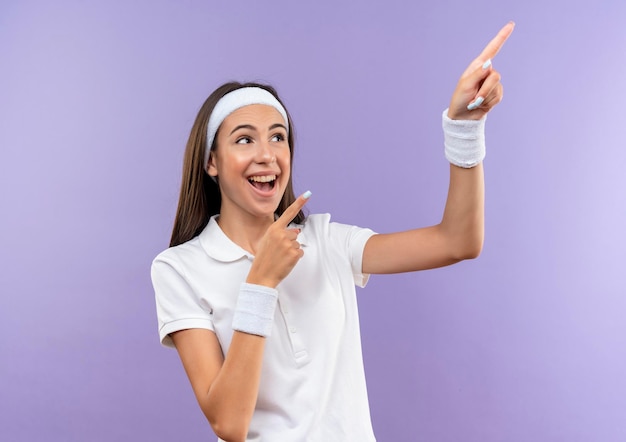 Joyeuse jolie fille sportive portant un bandeau et un bracelet pointant sur le côté isolé sur un mur violet