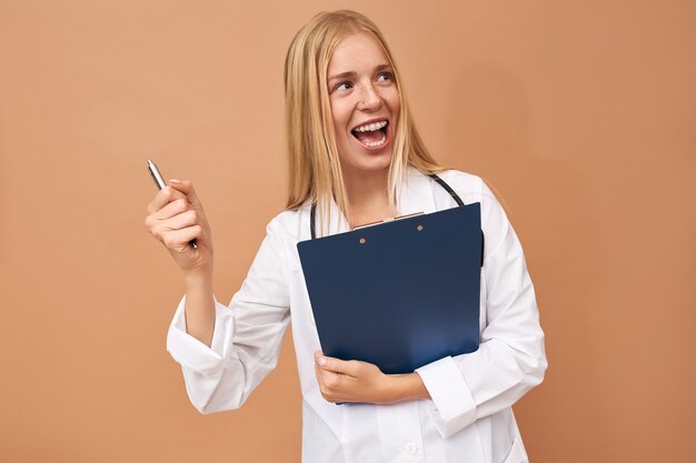 Joyeuse jolie jeune femme médecin en blouse chirurgicale blanche s'exclamant joyeusement avec la bouche grande ouverte, célébrant le succès tout en travaillant à l'hôpital, se réjouissant des bons résultats de tests sanguins