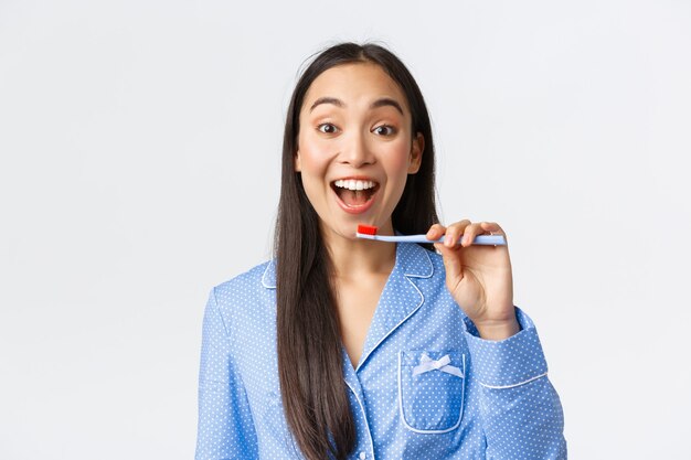 Joyeuse jolie jeune fille asiatique en pyjama bleu se réveillant, se brossant les dents avec un large sourire enthousiaste, tenant une brosse à dents près des dents blanches, fond blanc. Espace de copie