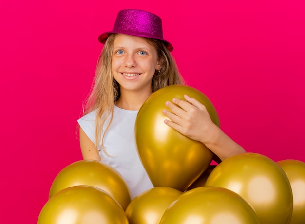 Photo gratuite joyeuse jolie petite fille en chapeau de vacances avec bouquet de ballons regardant la caméra en souriant avec un visage heureux, concept de fête d'anniversaire debout sur fond rose